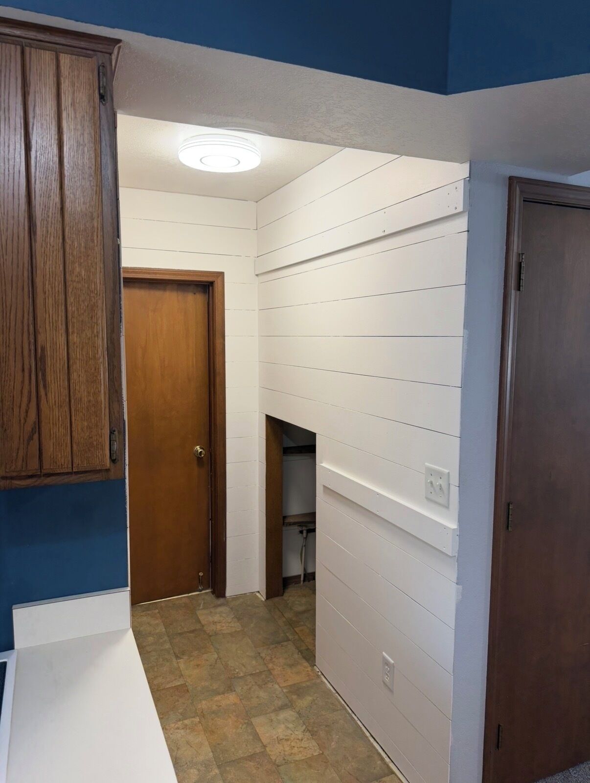 A narrow hallway with white paneling, a wooden door, and a ceiling light fixture.