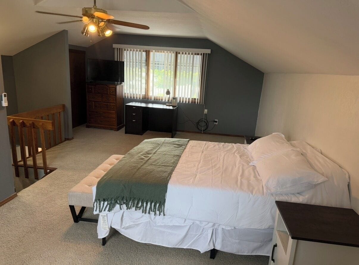 A bedroom with a bed, a desk, a chair, a television, and a window with blinds.