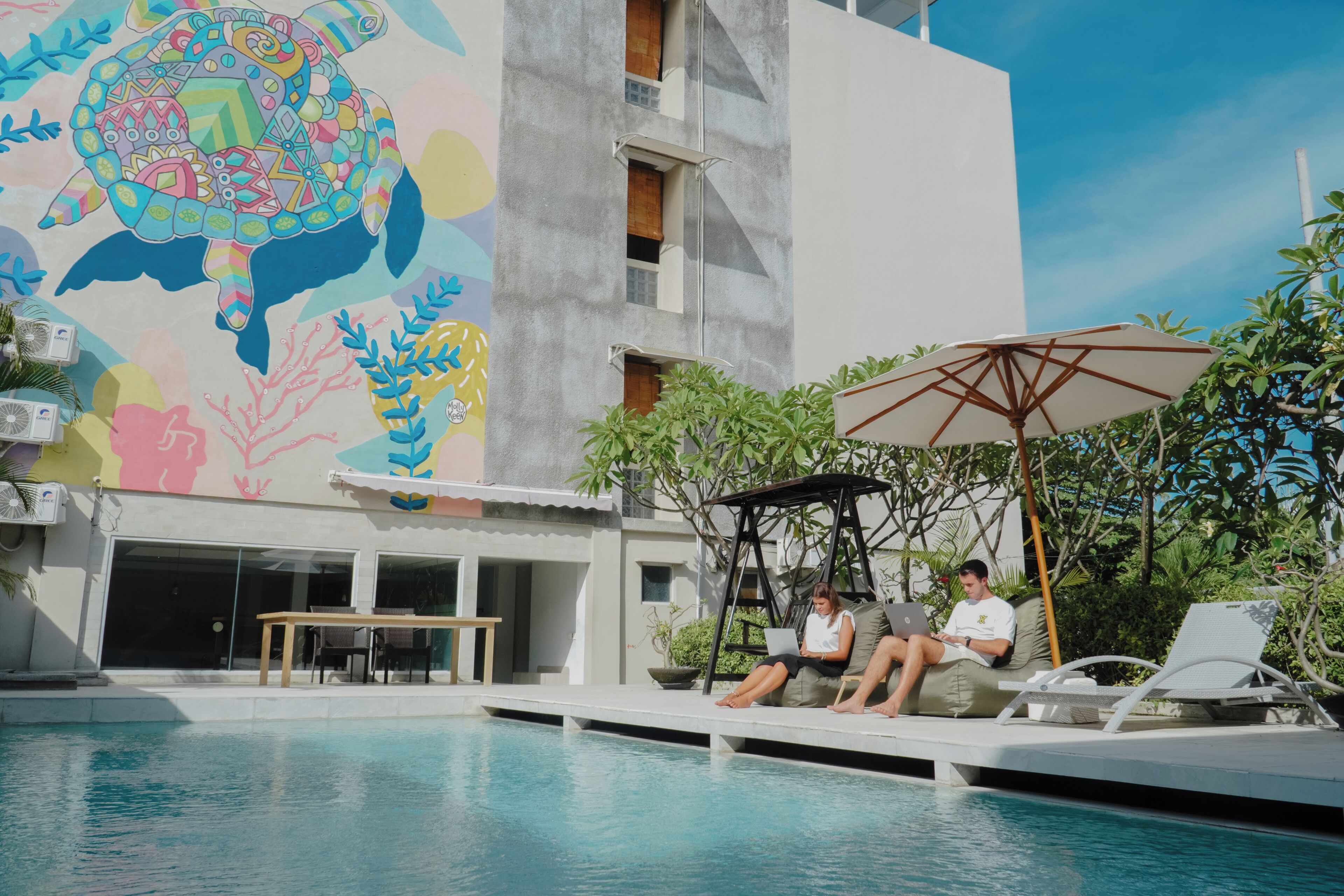 Una piscina con sillas de descanso y sombrillas, un mural en el edificio y un cielo despejado.