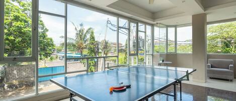 Una mesa de ping pong en una habitación con ventanas amplias, un ventilador de techo y un sofá.
