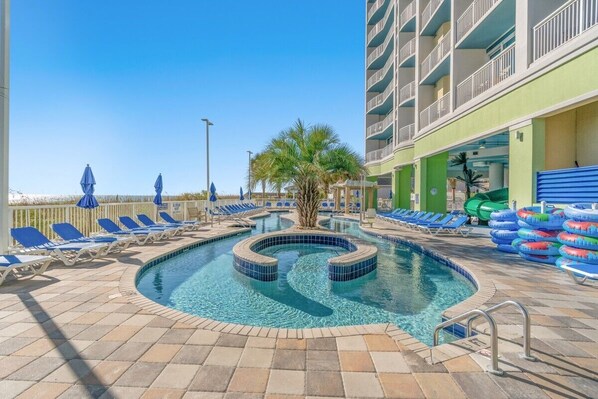 A swimming pool with lounge chairs and umbrellas, surrounded by a building with balconies.