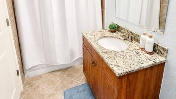 Un baño con cortina de ducha, un lavabo con encimera de mármol, un mueble de tocador de madera y un espejo.