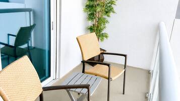 Un balcón con sillas de mimbre, una mesita, una planta en maceta y vistas del interior.