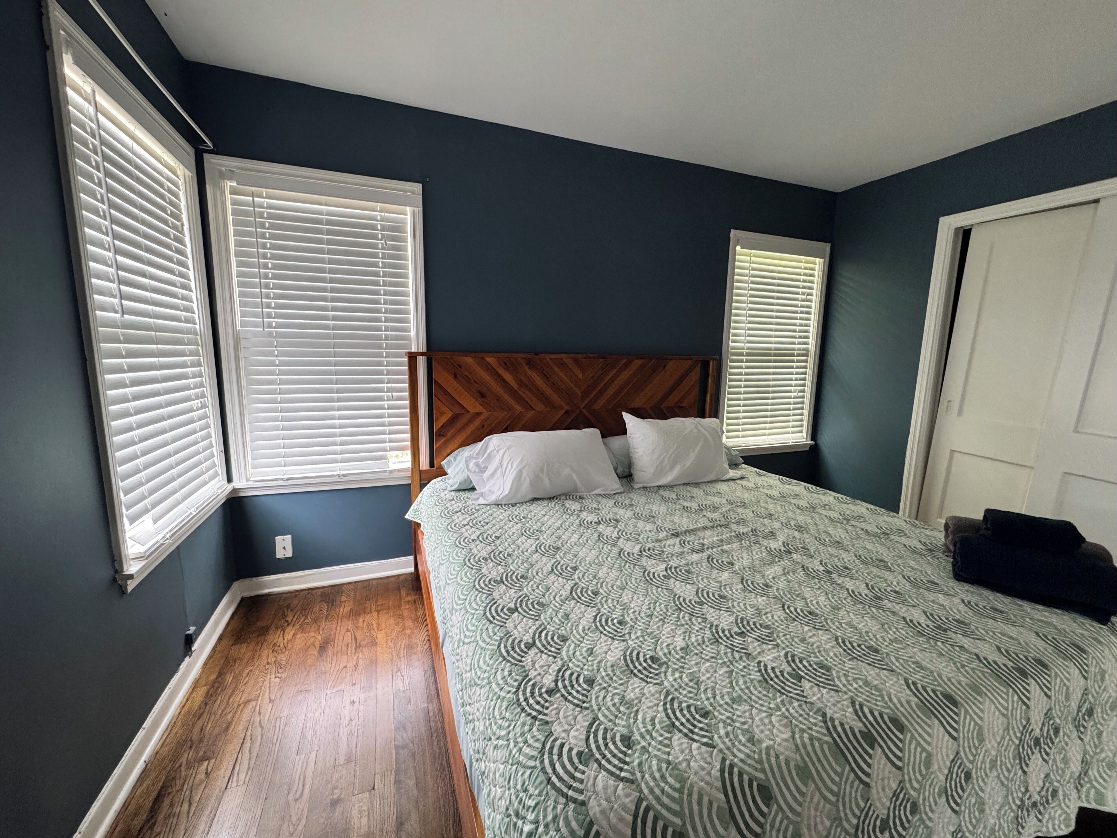 A bedroom with a large bed, wooden headboard, and three windows with blinds.