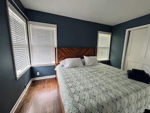 A bedroom with a large bed, wooden headboard, and three windows with blinds.