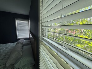 A bedroom with a bed, a window with blinds, and a view of greenery outside.