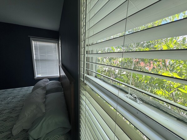 Un dormitorio con cama, ventana con persianas y vista a follaje exterior.