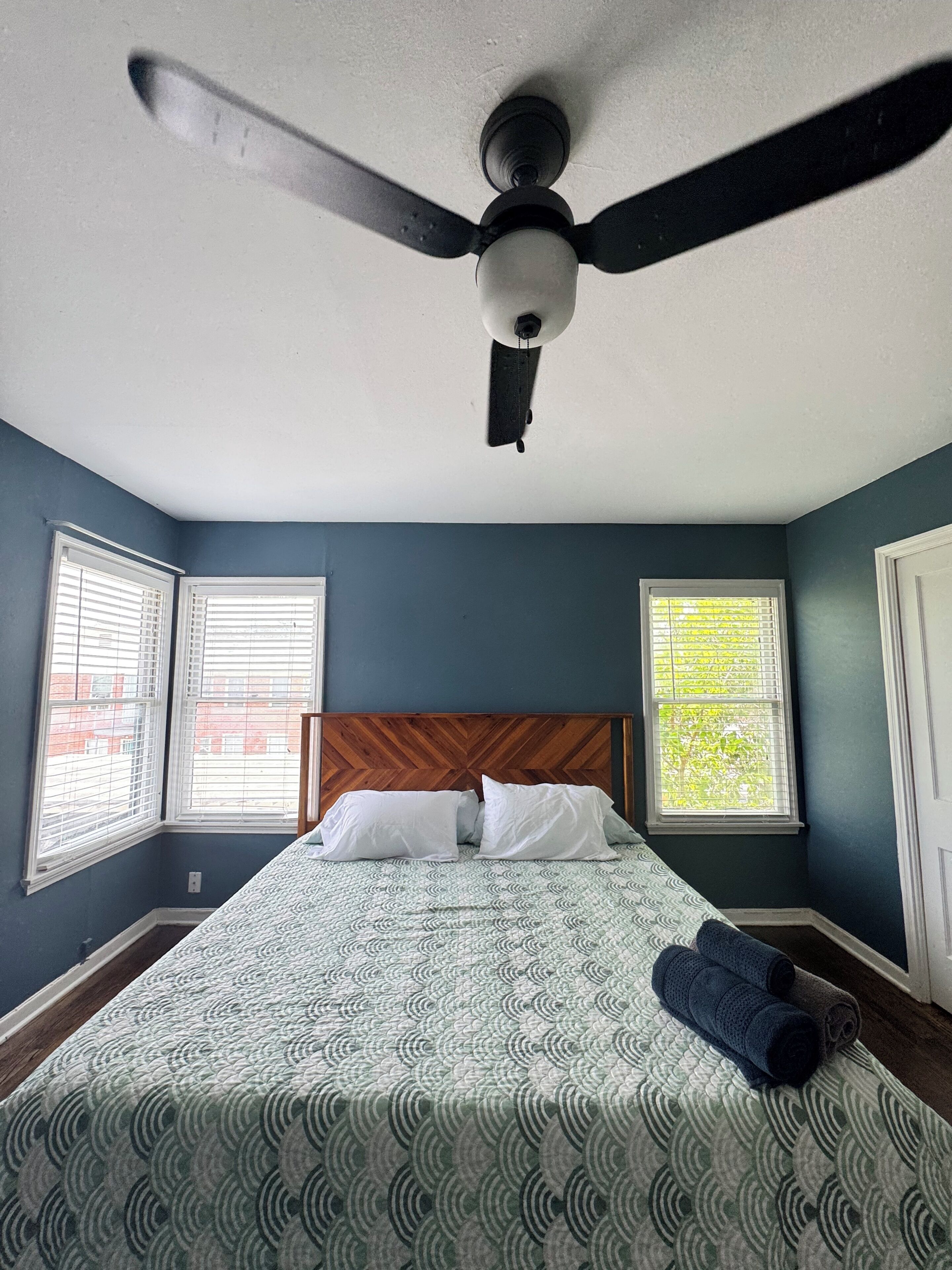 Ein Schlafzimmer mit einem Bett, zwei Jalousien an den Fenstern, einem Deckenventilator und einem hölzernen Kopfteil.