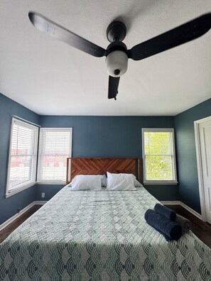 A bedroom with a bed, two windows with blinds, a ceiling fan, and a wooden headboard.