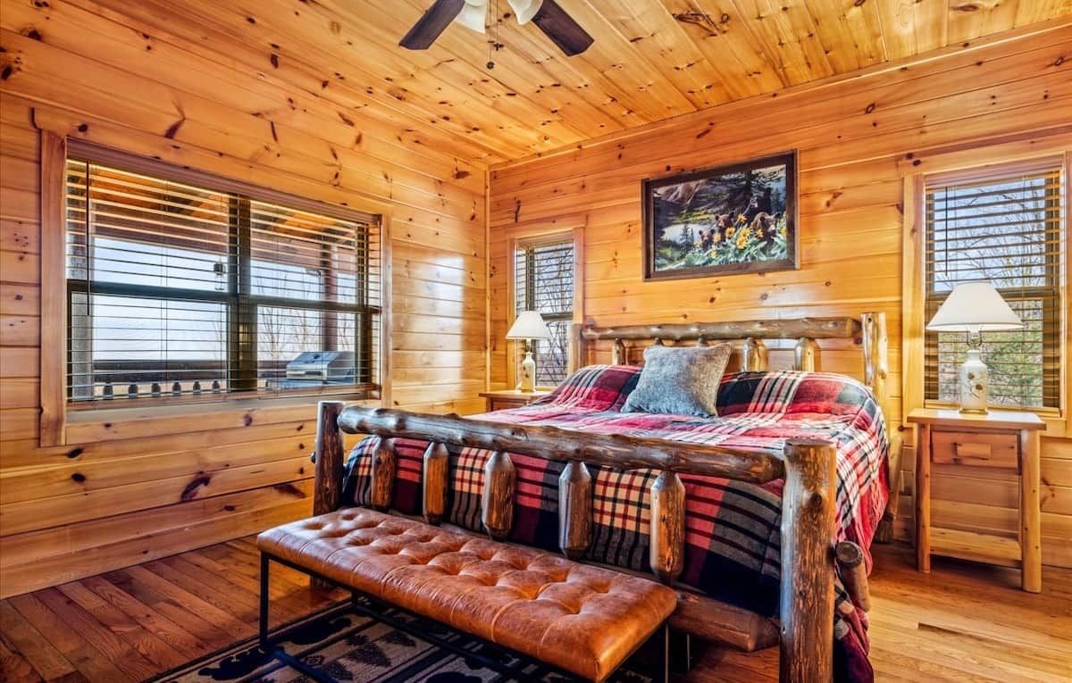 A log cabin bedroom with a bed, bench, and two windows with blinds.
