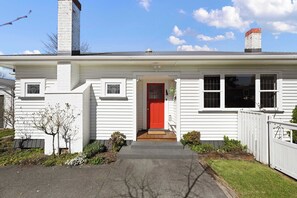 A single-story white house with a red front door, a chimney, and a small porch with a bench.