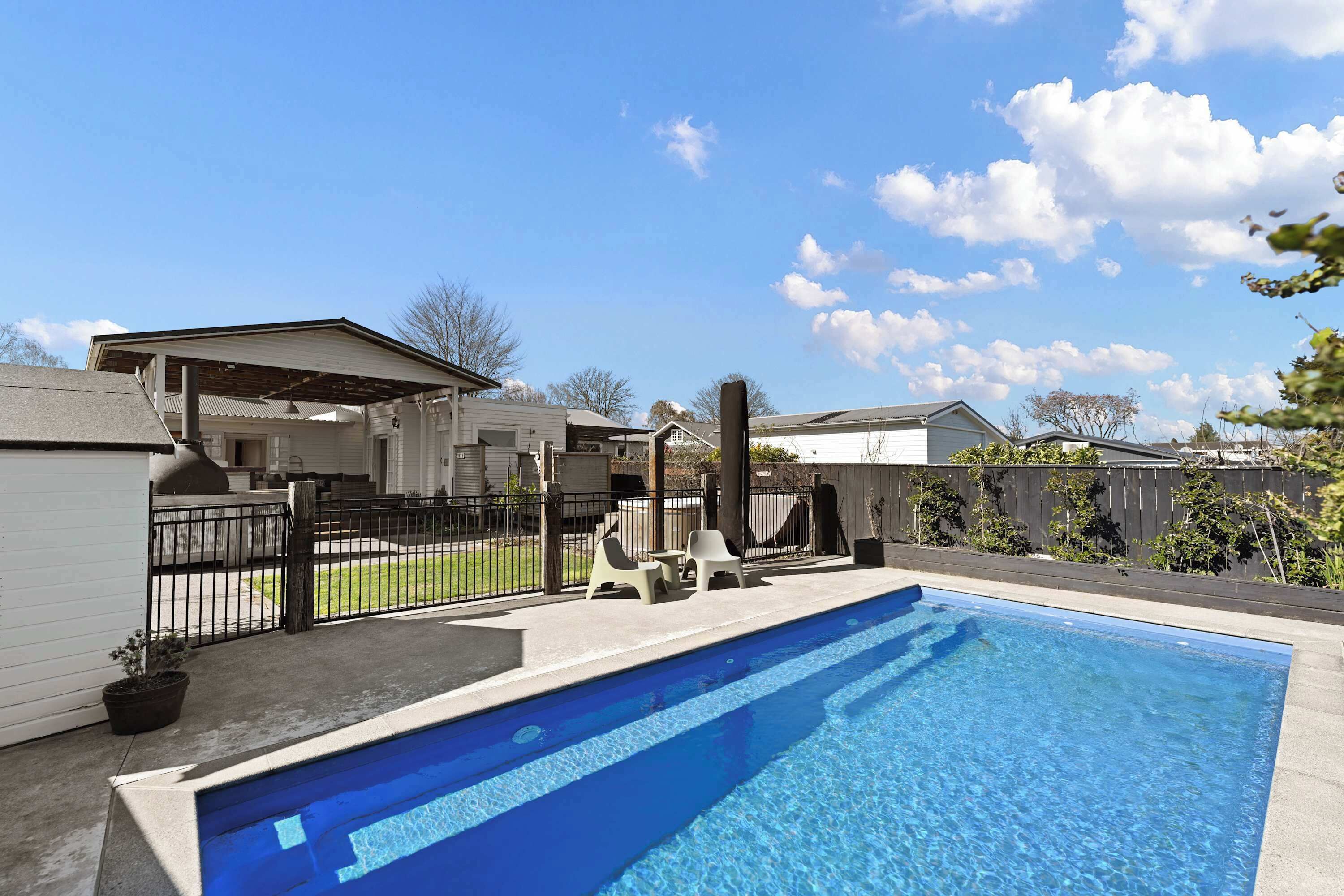 Una piscina residencial con un área de patio techada, un patio cercado y casas al fondo.
