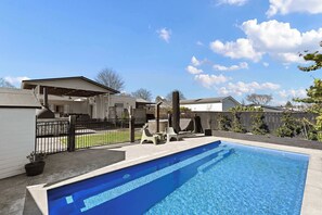 A residential swimming pool with a covered patio area, a fenced yard, and houses in the background.
