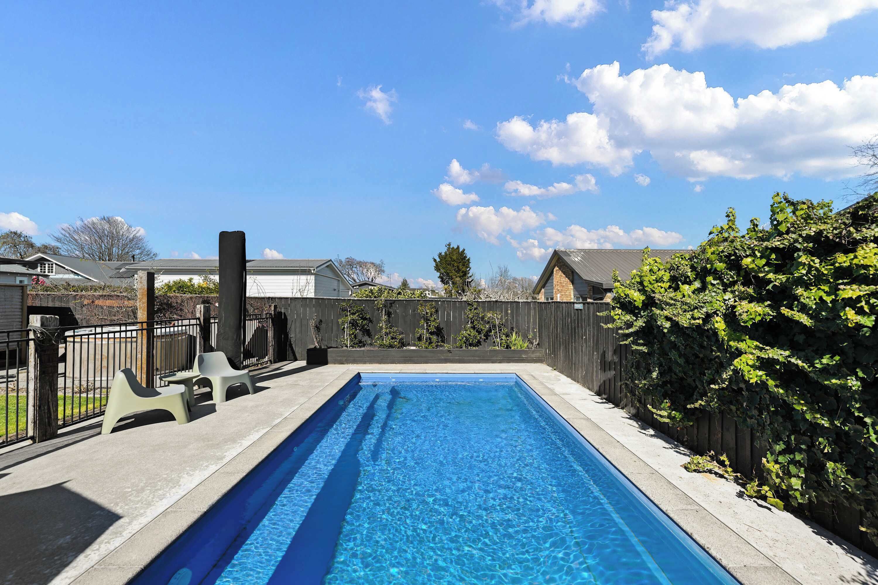 A rectangular swimming pool with clear blue water, surrounded by a concrete deck and a wooden fence.