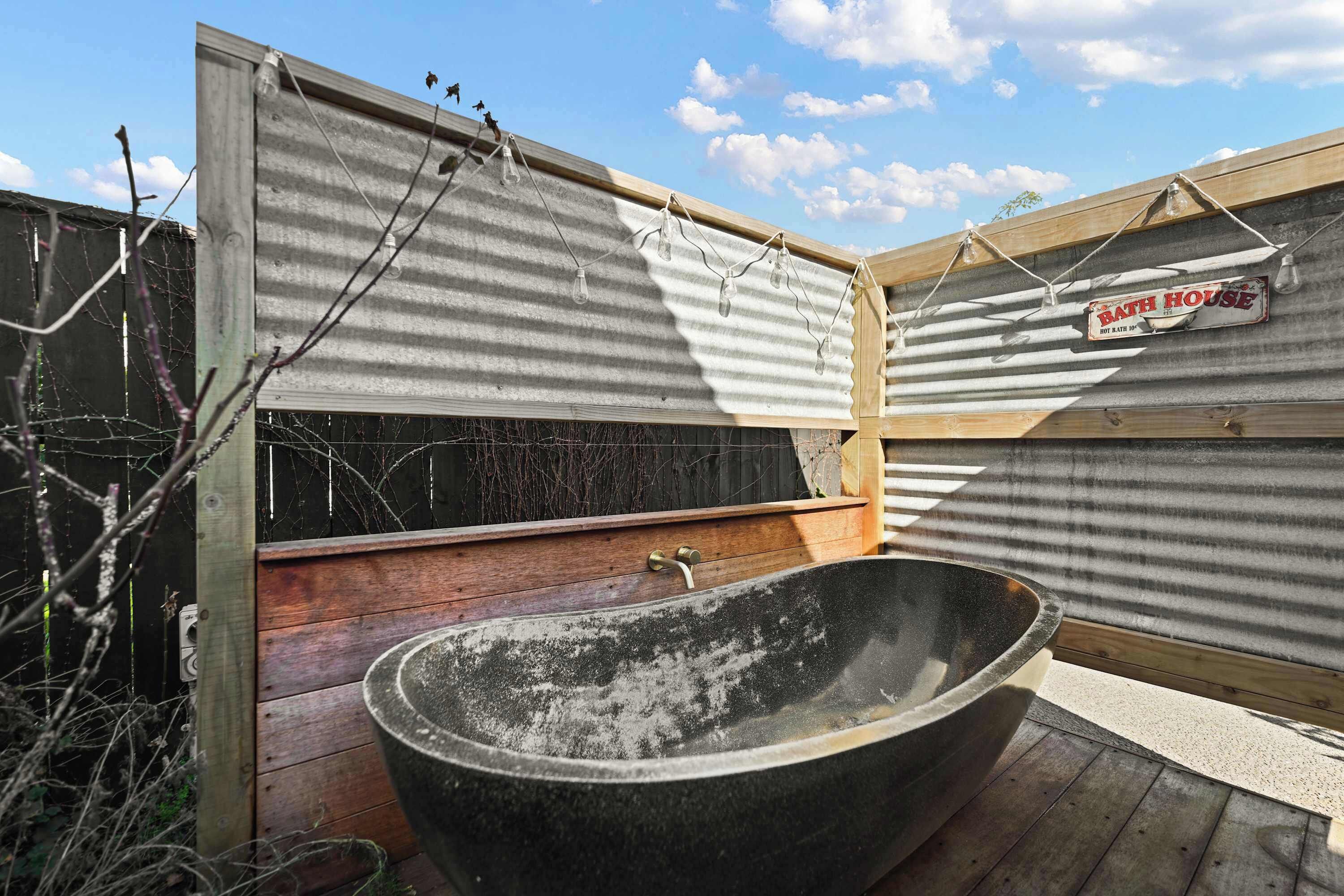 Ein rustikales Badezimmer im Freien mit freistehender Badewanne, Holzdeck und Wellblechwänden.