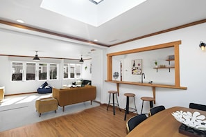 A modern living room with a wooden floor, a sofa, a dining table with chairs, and a large window with multiple panes.
