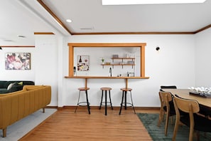 A modern living space with a wooden floor, a sofa, a dining table with chairs, and a kitchen area with stools and shelves.