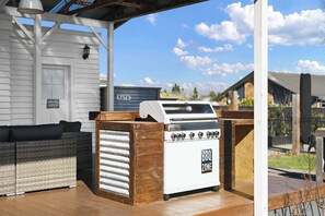 An outdoor barbecue grill with a 'BBQ Zone' sign, a wicker sofa, and a covered patio area with a 'Bath' sign in the background.