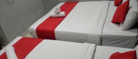 Duas camas de solteiro com roupa de cama vermelha e branca, uma televisão de tela plana fixada na parede e uma tomada elétrica à esquerda.