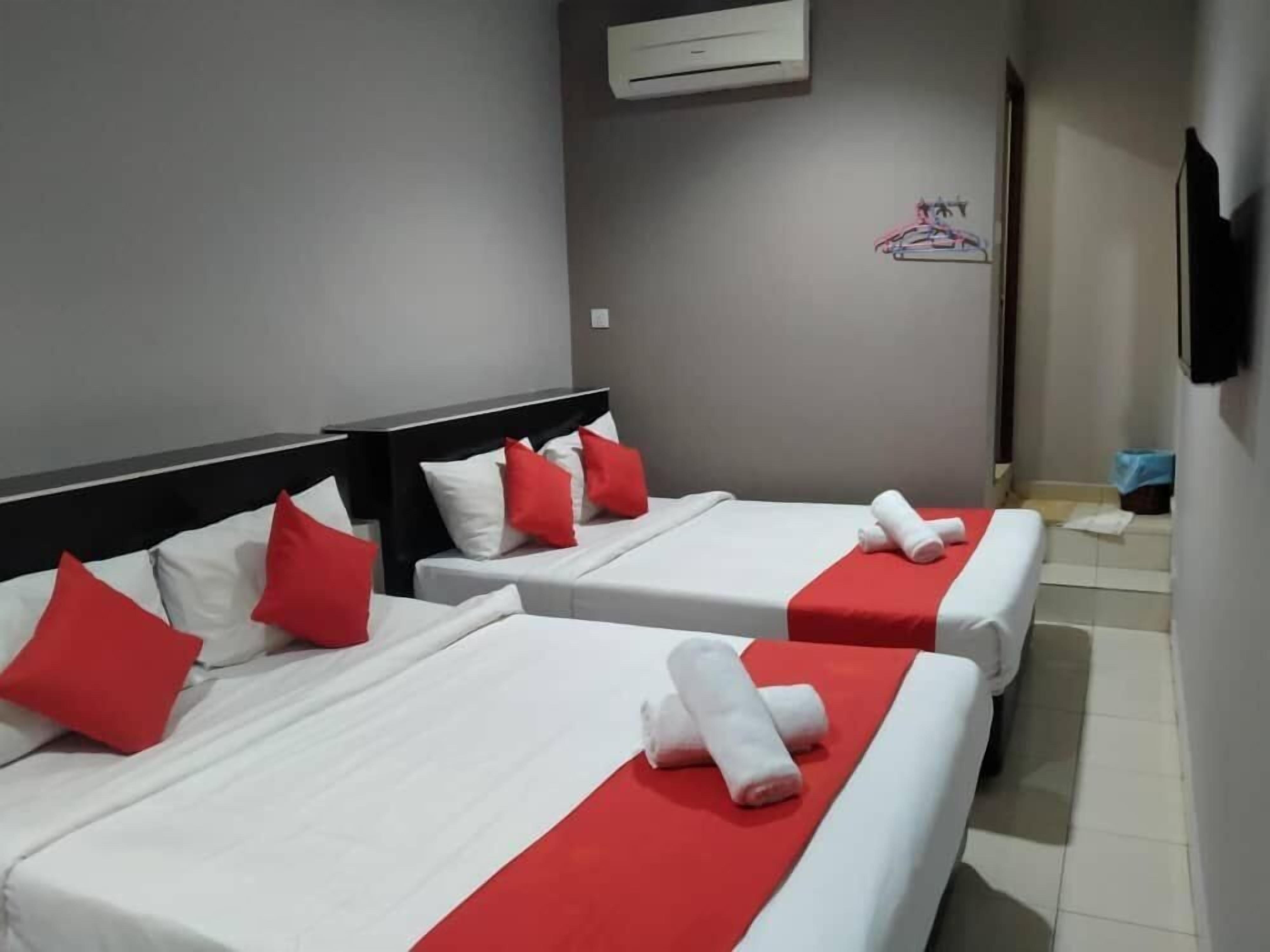 Habitación de hotel con dos camas, cada una con ropa blanca y detalles rojos, un televisor de pantalla plana en la pared y un equipo de aire acondicionado sobre las camas.