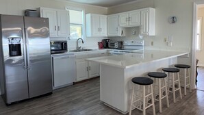 Una cocina moderna con armarios blancos, electrodomésticos de acero inoxidable, una isla central con taburetes y una ventana con persianas.