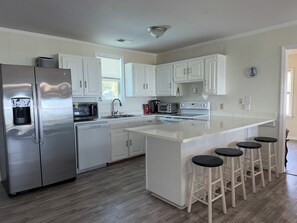 Una cocina moderna con armarios blancos, electrodomésticos de acero inoxidable, una isla central con taburetes y una ventana con persianas.