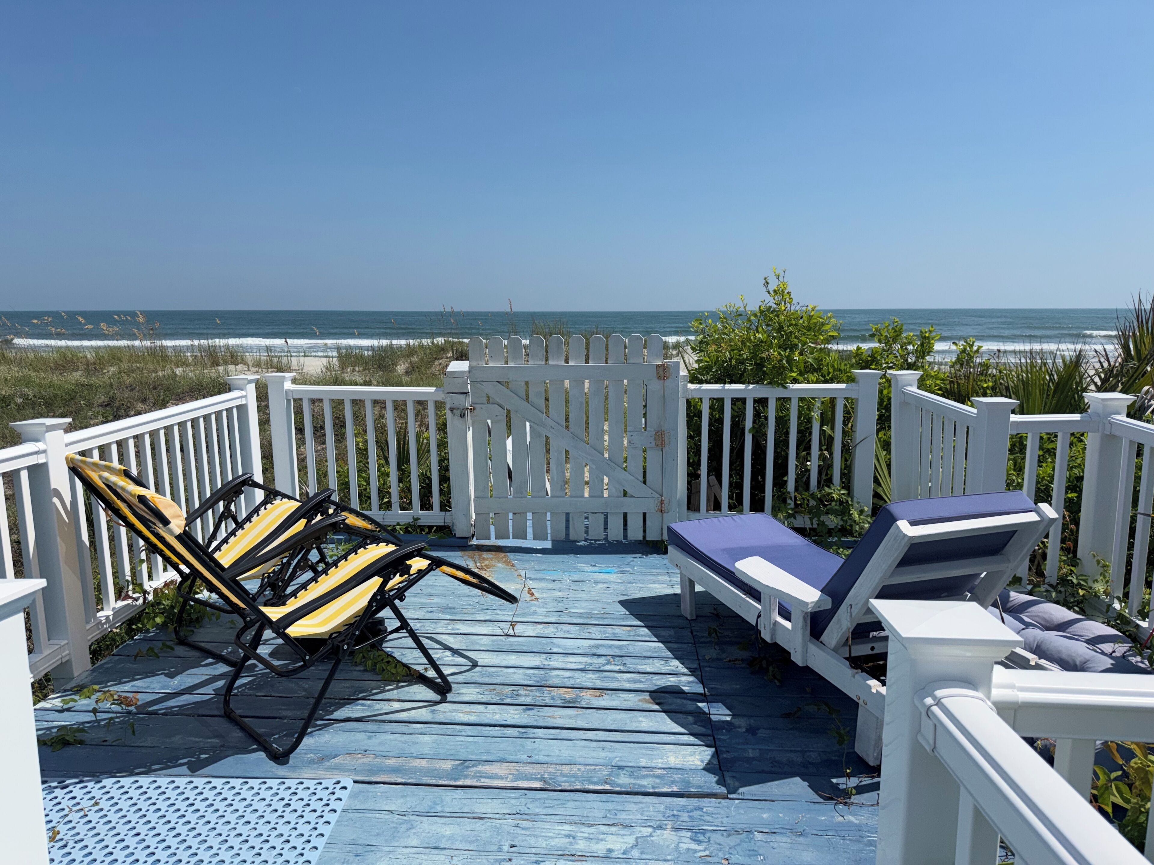 A wooden deck with two sun loungers and a gate, overlooking a beach.