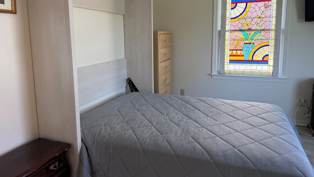 Un dormitorio con cama, cómoda, armario y una ventana con vitrales.