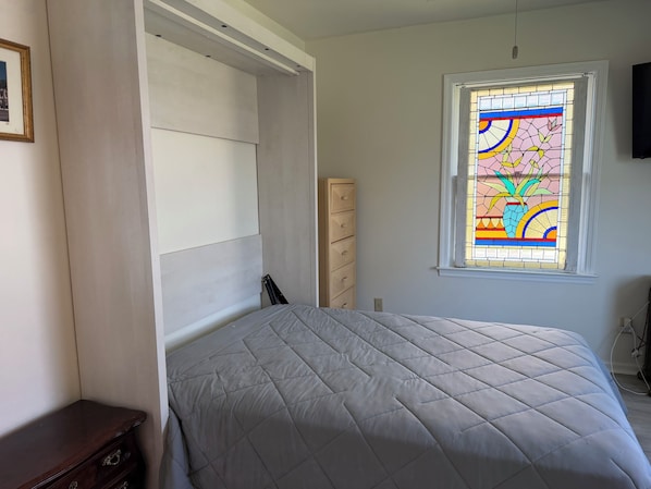 Un dormitorio con cama, cómoda, armario y una ventana con vitrales.