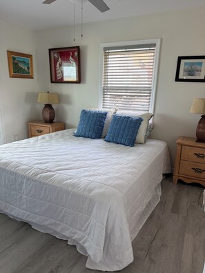 A bedroom with a large bed, two bedside tables, a window with blinds, and wall-mounted artwork.
