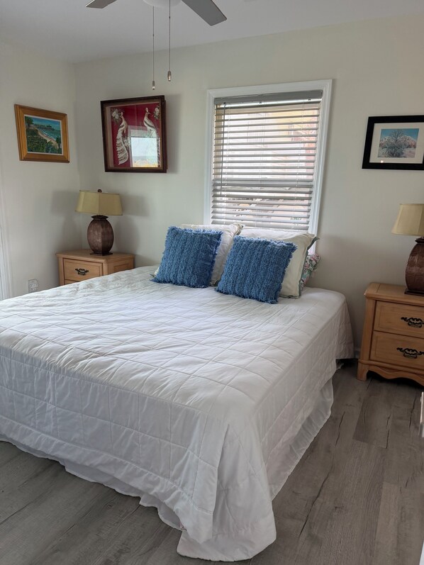 A bedroom with a large bed, two bedside tables, a window with blinds, and wall-mounted artwork.