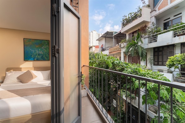 A hotel room with a bed, a painting on the wall, and a balcony with a view of neighboring buildings and greenery.