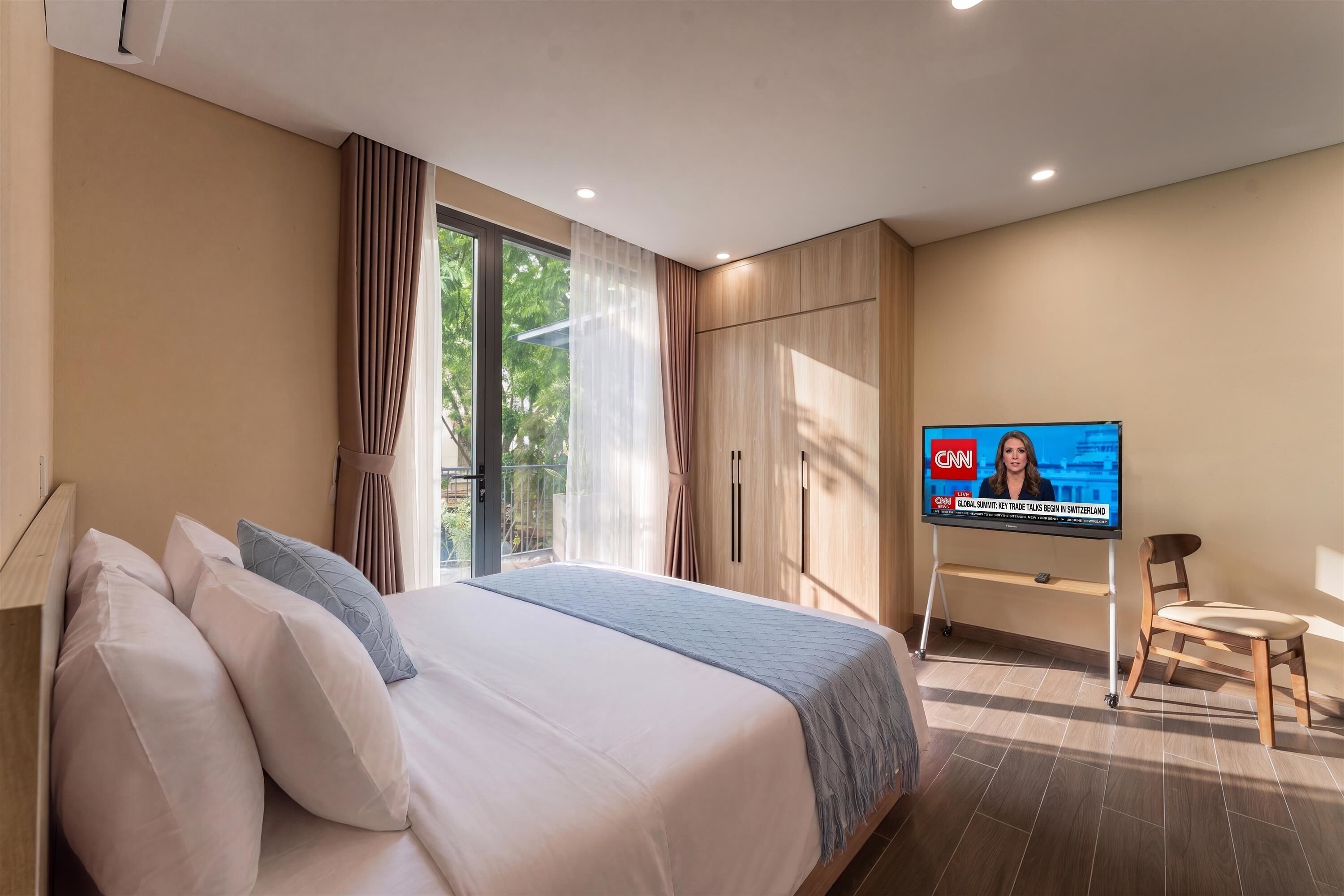 A hotel room with a large bed, a television displaying a news program, and a view of greenery outside.