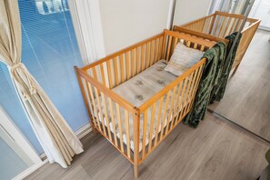 A wooden crib with a mattress and pillows, positioned next to a window with blue blinds.