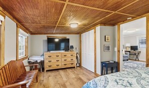 A room with a wooden ceiling, a bed with a patterned bedspread, a wooden dresser, a television, and a window.