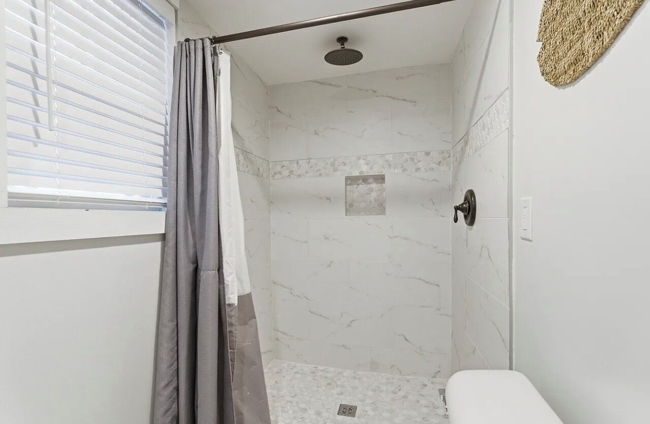 A bathroom with a shower, a toilet, and a window with blinds.
