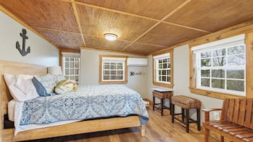 A bedroom with a wooden ceiling, a bed with patterned bedding, two windows, a small table, and a bench.