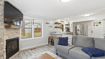 Uma sala de estar moderna com lareira, sofá cinzento e área de cozinha com prateleiras de madeira.