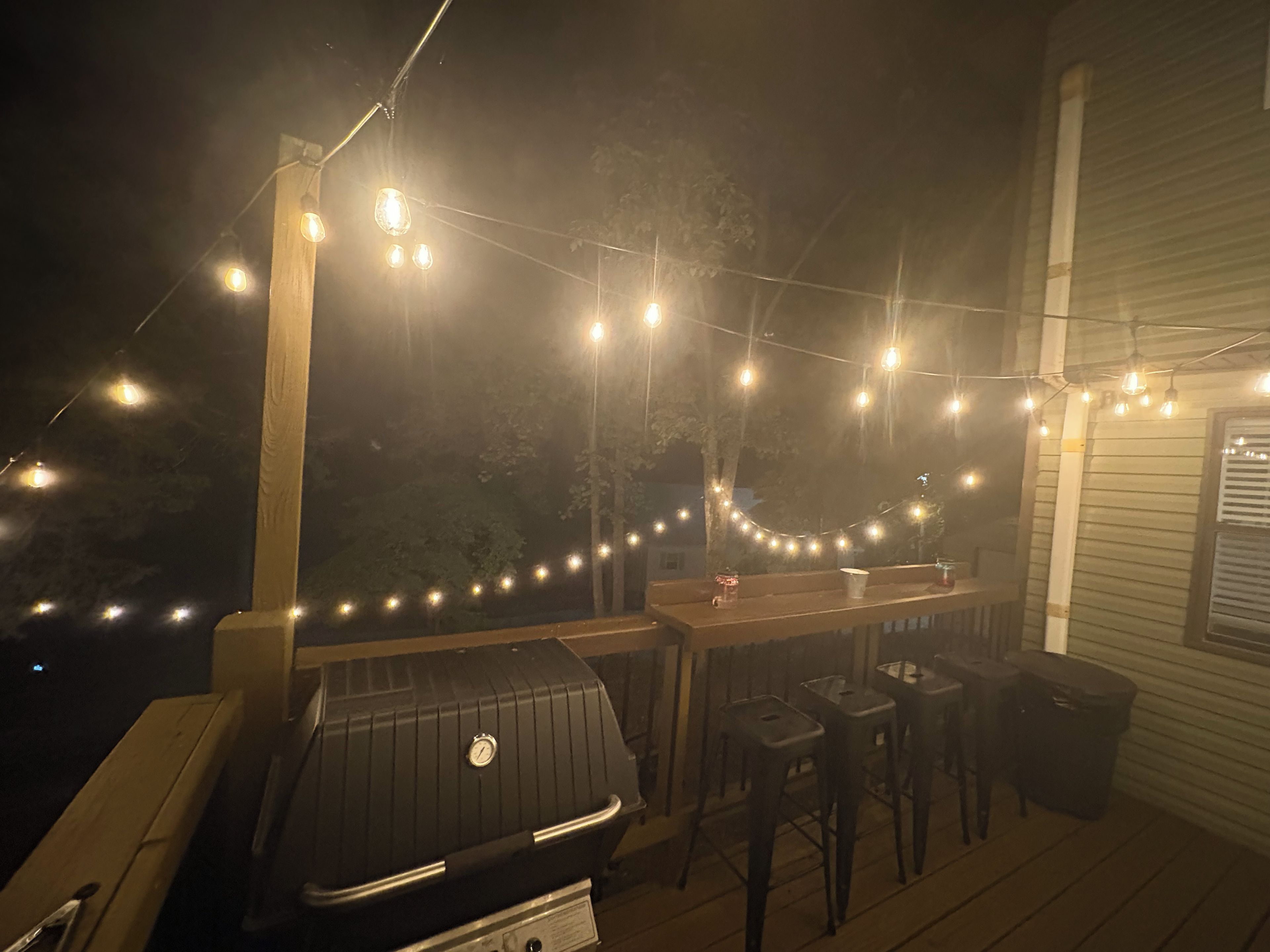 An outdoor deck with a barbecue grill, wooden chairs, and string lights.