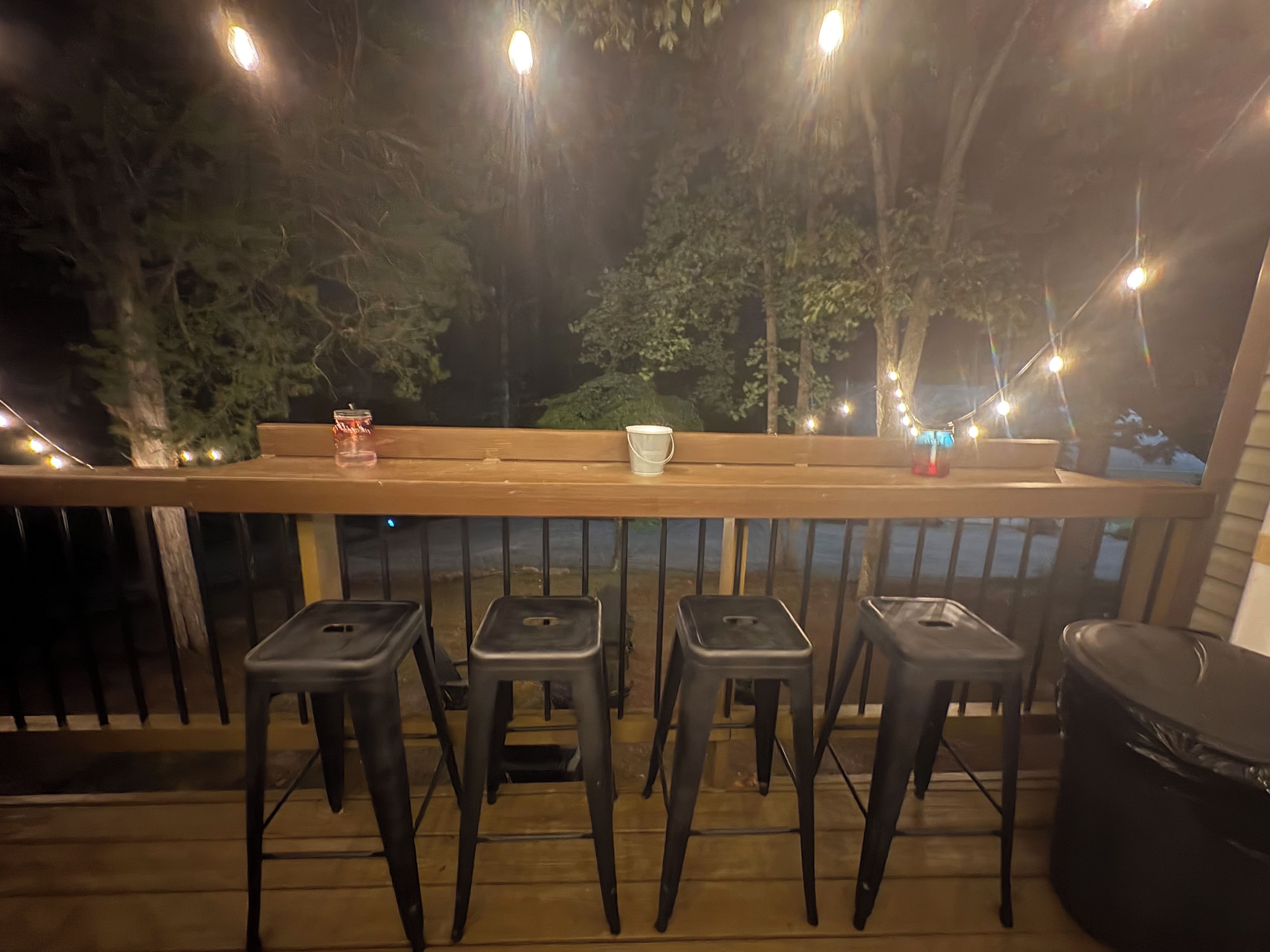 A wooden deck with a railing, four black stools, a bench, and string lights.