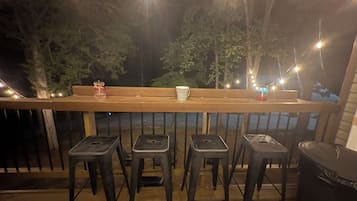 A wooden deck with a railing, four black stools, a bench, and string lights.