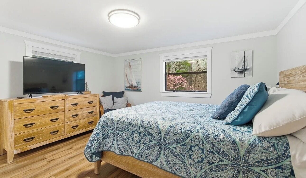 A bedroom with a bed, a TV, a wooden dresser, and a window with a view of trees.