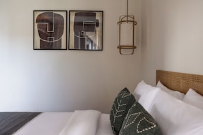 Een bed met witte lakens, een geweven hoofdbord en twee ingelijste abstracte kunstwerken aan de muur.