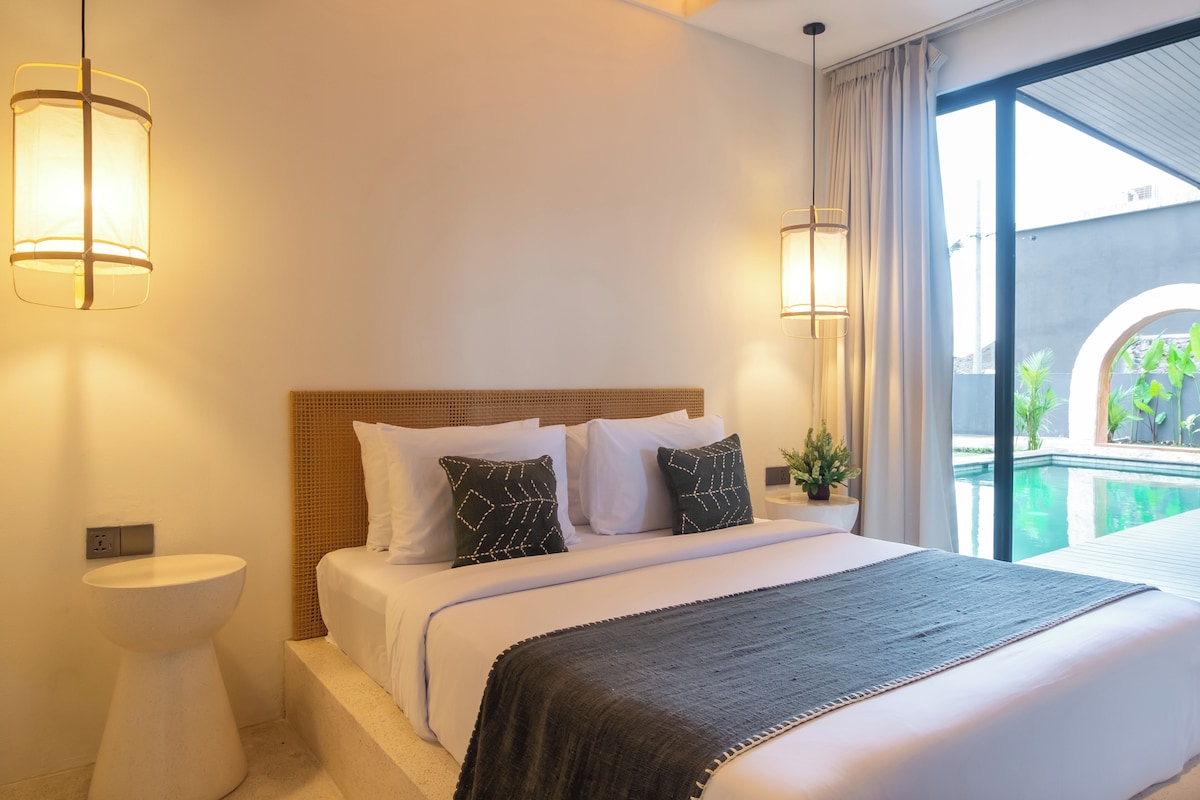 A hotel room with a bed, bedside tables, a window with a view of a pool and greenery, and wall-mounted lamps.