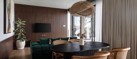 A modern living room with a round dining table, green sofa, wicker chairs, a large potted plant, and a flat-screen TV.