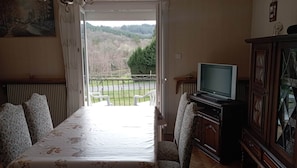 A dining area with a table and chairs, a television on a stand, and a view of a forested landscape through an open door.