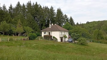A white house with a brown roof, surrounded by a grassy field and dense forest.