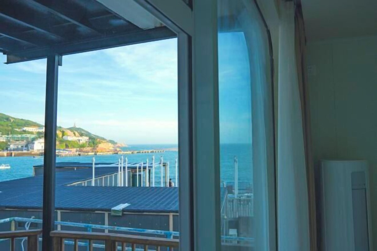 A view from a room with a large window overlooking a harbor, a pier, and a distant coastline.