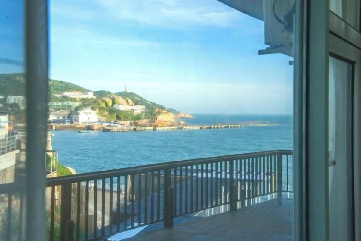A balcony with a view of the sea, a lighthouse, and buildings on a hill.