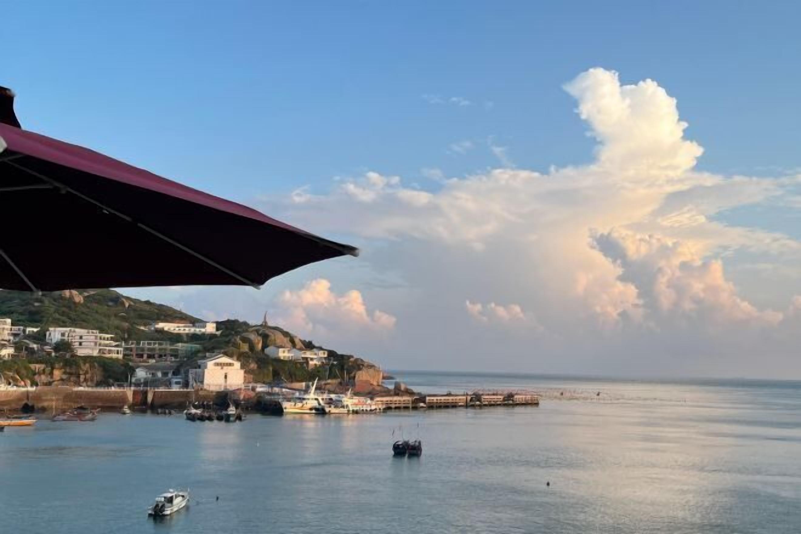 Vista costera con un muelle que se adentra en aguas tranquilas, cielo despejado y un edificio a la izquierda.